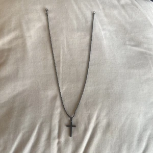 Svart halsband med korshänge - Stilrent svart halsband med ett korsformat hänge. Kedjan är tvinnad i nytt skick, kedjan är gjord av stål, 50cm lång, 75kr