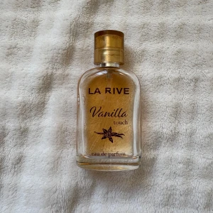 La Rive Vanilla Touch parfym - En söt och varm parfym från La Rive med tydlig vaniljdoft. Flaskan är genomskinlig med rundade former och har en gyllene kork. Innehållet är ljusgult och flaskan pryds av bruna detaljer och en vaniljblomma på framsidan.