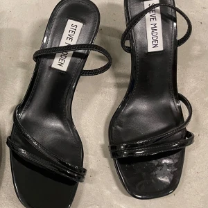 Svarta sandaler med klack från Steve Madden - Stilrena svarta sandaler från Steve Madden med smala remmar och fyrkantig tå. Skorna har en elegant låg klack och är tillverkade i glansigt syntetmaterial. Perfekta för dig som gillar minimalistisk och trendig design.