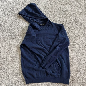 Mörkblå hoodie från JAJA i ullmix - Mörkblå hoodie från JAJA i en skön mix av ull, bomull och spandex. Klassisk modell med huva och långa ärmar, perfekt för kyliga dagar. Enkel och stilren design utan tryck eller mönster, passar dig som gillar basic och comfy vibes. 