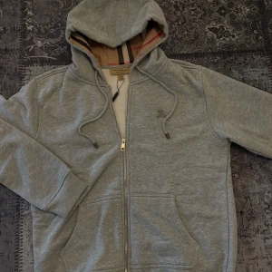 Grå hoodie från Burberry med dragkedja - Snygg grå hoodie från Burberry, helt ny och aldrig använd. Pris kan diskuteras. Skickas samma dag ✅ Ställ gärna frågor eller funderingar!