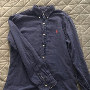 Blå skjorta från Polo Ralph Lauren - Klassisk marinblå skjorta från Polo Ralph Lauren i slim fit-modell. Skjortan har långa ärmar, knappar framtill och den ikoniska röda logotypen broderad på bröstet. Tillverkad i mjuk bomull och passar perfekt till jeans eller chinos.