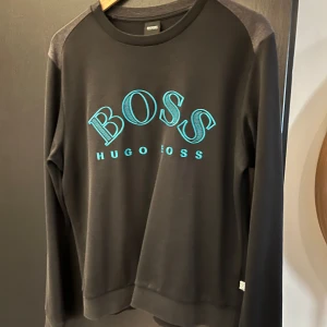 Hugo Boss sweatshirt  - Stilren sweatshirt från Hugo Boss. Logan och söm på ryggen är i ljusblå neon färg. Storleken på tröjan är M. Den har använts lite men är fortfarande i bra skick( finns det litet hål på ryggen se bild 5 )