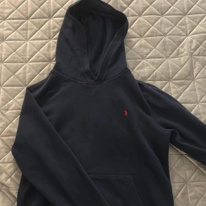 Mörkblå hoodie från Polo Ralph Lauren - Snygg mörkblå hoodie från Polo Ralph Lauren med klassisk röd logga broderad på bröstet. Tröjan har huva, lång ärm och en stor magficka framtill. Perfekt för dig som gillar stilren och enkel streetwear.