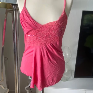 Rosa linnetopp med spets och paljetter - Supersöt rosa linnetopp med smala axelband och omlott-detalj framtill. Toppen har ett parti med spets och glittrande paljetter över bysten som ger extra wow-känsla. Mjuk och lätt i materialet, perfekt för varma dagar eller när du vill sticka ut lite extra. Storlek S