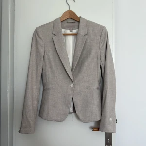 Beige kavaj från HM - Beige kortare kavaj från HM. Kavajen har intagen midja, klassisk krage, en knapp framtill och två fickor. Tunna axelvaddar. Materialet är lätt strukturerat.