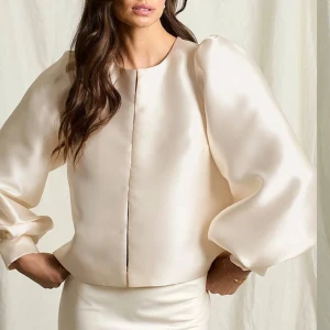 Champagnefärgad satinblus - Supersnygg champagnefärgad med puffiga långärmar och rund halsringning. Jackan har knappar framtill och en lyxig, glansig finish som verkligen sticker ut. Perfekt för dig som vill ha något unikt och trendigt.