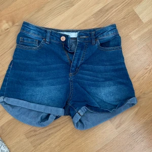 Blå jeansshorts  - Snygga blå jeansshorts från Amisu med klassisk femficksdesign, uppvikta benslut och bälteshällor. Perfekta för sommaren med sin mörkblå tvätt och bekväma passform. Stängs med knapp och dragkedja framtill. Högmidjade. 