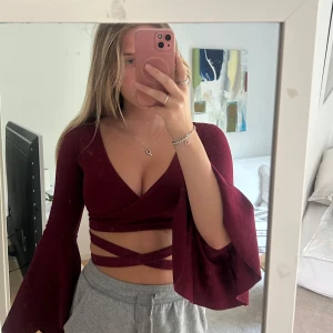 Vinröd omlottopp med utsvängda ärmar - Snygg vinröd omlottopp med djup v-ringning och långa utsvängda ärmar. Toppen är croppad och har trendiga snörningsdetaljer runt midjan som ger en cool look. Perfekt för dig som vill sticka ut med en unik och stilren topp.