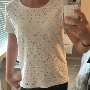 Vit spetsblus från Jacqueline de Yong - Superfin vit blus från Jacqueline de Yong i storlek S. Toppen har korta ärmar och är gjord i ett spetsliknande bomullstyg med snyggt hålmönster i bladform. Perfekt för dig som gillar en luftig och stilren look med lite extra detalj.