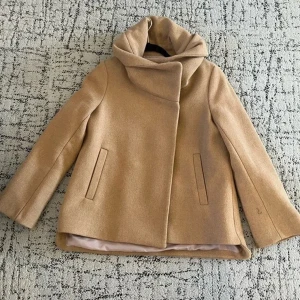 Beige ullkappa med stor krage och luva - Snygg beige kappa i ull med oversized krage som ger en cool och modern look. 