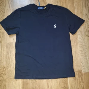 Svart t-shirt från Polo Ralph Lauren - S men kan passa M. Klassisk svart t-shirt från Polo Ralph Lauren med vit broderad logga på bröstet. T-shirten har rund halsringning och korta ärmar. Tillverkad i mjuk bomull för en skön känsla och stilren look.