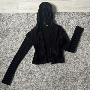 Svart ribbad hoodie från H&M Divided - Svart ribbstickad hoodie från H&M Divided med dragkedja framtill och långa ärmar. Tröjan har huva med snörning och är i en tight passform. Perfekt för dig som gillar en enkel men ändå snygg look.