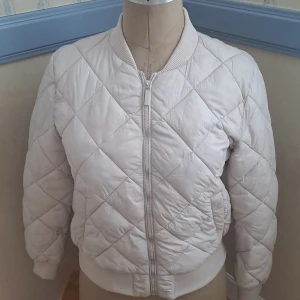 Beige quiltad bomberjacka - Beige bomberjacka från Åhléns i storlek M. Jackan är quiltad med rutmönster, har dragkedja framtill och ribbade muddar vid hals, ärmslut och nederkant. Använd sparsamt. 🩷 str. 40/42