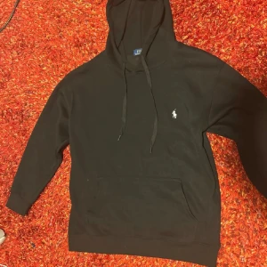 Svart hoodie från Polo Ralph Lauren - Svart hoodie från Polo Ralph Lauren med klassisk huva och snörning. Tröjan har en stor magficka och den ikoniska lilla broderade loggan på bröstet. Perfekt för en chill och stilren look. Tillverkad i mjuk bomull för extra komfort. Funkar som S också 