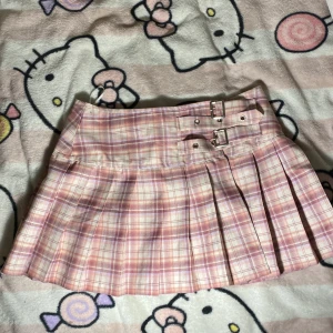 Rutig rosa kortkjol från Banned Alternative - Supersöt kortkjol i rosa och beige rutigt från Banned Alternative. Kjolen har omlottmodell med två snygga spännen i midjan och fina veck. Perfekt för dig som gillar en preppy eller alternativ stil!