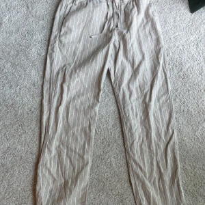 Beige randiga linnebyxor med snörning - Snygga beige linnebyxor med vita ränder och avslappnad passform. Byxorna har elastisk midja med snörning och raka ben, perfekta för en chill och stilren look. Lätta och luftiga, passar grymt till sommaren. Linnebyxorns är från HM i storlek S kontakta vid minsta fundering 
