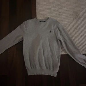 Grå stickad tröja Polo Ralph Lauren - Säljer en stilren grå stickad tröja från Polo Ralph Lauren. Tröjan har rund halsringning, diskret svart logga broderad på bröstet och ribbade muddar vid ärmslut och nederkant. Perfekt för lager-på-lager och enkel att matcha.