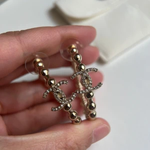Chanell örhängen med pärldetaljer - Snygga hoop-örhängen i guldton med pärlformade kulor och glittrande CC-logga täckt av små stenar. Statement-accessoar som verkligen drar blickarna till sig. Perfekta för dig som vill ha något extra och älskar lyxiga detaljer.