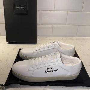 Vita Saint Laurent sneakers i skinn - Snygga vita sneakers i slät skinn med logga på sidan. Klassisk låg modell med rund tå, vita snören och diskret guldig text på plösen. Perfekta för dig som gillar stilrena och exklusiva skor.