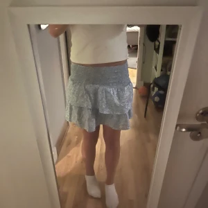 Ljusblå volangkjol med blommigt mönster - Supersöt ljusblå kortkjol med små vita blommor och volangdetaljer. Kjolen har bred smock i midjan som gör den bekväm och sitter snyggt. Perfekt för dig som gillar en feminin och somrig stil!