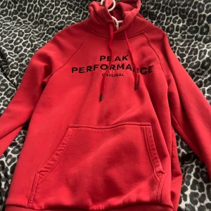 Röd hoodie Peak Performance - Röd hoodie från Peak Performance med svart broderad logga på bröstet. Tröjan har huva med snörning och en stor magficka. Perfekt för dig som gillar en sportig och avslappnad stil.