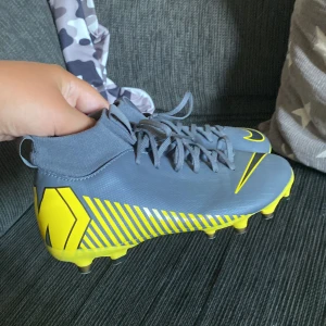 Nike grå och gula fotbollsskor - Snygga fotbollsskor från Nike i grått med gula detaljer och coola grafiska mönster på sidorna. Skorna har snörning, högre strumpliknande skaft och plös, samt platt sula med dobbar för bra grepp på planen. Perfekta för dig som vill sticka ut på matchen.