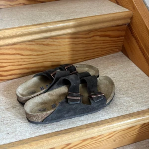 Svarta mockasandaler från Birkenstock storlek 39 passar även 40 - Klassiska svarta sandaler från Birkenstock i mocka med två breda remmar och justerbara metallspännen. Fotbädden är i ljus mocka och sulan är svart med vågigt mönster. Perfekta för sommaren och riktigt sköna att gå i.