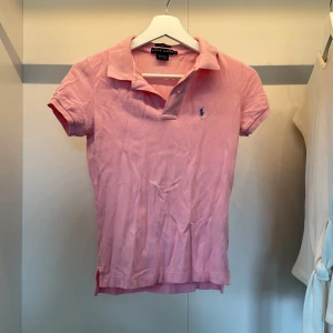 Rosa pikétröja från Ralph Lauren XS - Snygg rosa pikétröja från Ralph Lauren i klassisk modell med krage och knappar framtill. Tröjan har korta ärmar och den ikoniska blå loggan broderad på bröstet. Tillverkad i 100% bomull för en skön och luftig känsla.