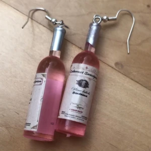 Örhängen med vinflaskor i rosa - Unika örhängen formade som små vinflaskor med rosa vätska och detaljerad etikett. Flaskorna har silverfärgade korkar och krokar. Perfekt accessoar för dig som vill sticka ut och visa din kärlek till vin! Priset gäller för ett par örhängen (som visas på bilden) men finns 4 par i lager (de i bilden inkluderade) så man kan köpa alla fyra paren för 30kr.💞