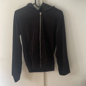 Svart velour hoodie från 157 XS - Mjuk och skön svart hoodie i velour från 157. Tröjan har huva, långa ärmar och en praktisk dragkedja framtill. Känguruficka på magen och ribbade muddar vid ärmslut och nederkant. Perfekt för en chill och avslappnad stil.
