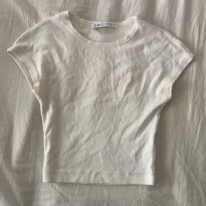 Vit croppad topp från Zara - Enkel och stilren vit croppad topp från Zara i mjuk bomull. Modellen har rund halsringning och korta ärmar med en något loose passform. Perfekt att matcha med jeans eller kjol för en clean look.