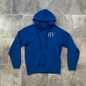 Blå hoodie från Bvaldi med strass - Blå hoodie från Bvaldi med dragkedja och huva. Framsidan har VB i strass och på ryggen står det BVALDI i stora bokstäver med både strass och blått tryck. Mjuk bomullskänsla och känguruficka framtill. Perfekt för dig som gillar streetstyle.