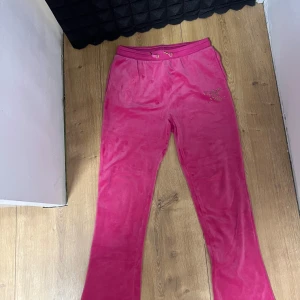 Juicy Couture rosa mjukisbyxor - Säljer ett par ikoniska rosa mjukisbyxor från Juicy Couture i mjuk velour. Byxorna har snörning i midjan, raka ben och bakfickor. På framsidan finns Juicy-loggan i strass för extra bling. Perfekta för en chill och trendig look. Har bara haft på mig de som pyjamas så är knappt använda 