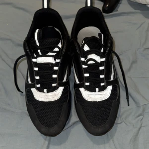 Svarta Dior sneakers med vita detaljer - Chunky svarta Dior sneakers med vita detaljer och reflekterande inslag. Ovandel i mesh och skinn, med kraftig sula och snörning. Snygg sportig vibe och tydlig Dior-logga på sidan. Perfekta för dig som gillar statement-sneakers med exklusiv känsla.