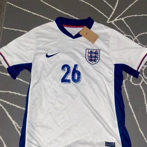 England Mainoo 26 Nike fotbollströja - England fotbollströja med Mainoo och nummer 26. Vit med blå detaljer, blå Nike-logga och landslagsemblem på bröstet. Kortärmad modell med blå sidor och krage, samt färgade ränder på ärmsluten. Tillverkad i lätt och ventilerande material.