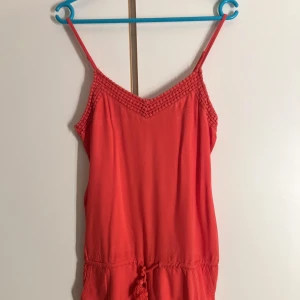 Playsuit - Superfin korallrosa playsuit från ONLY med smala axelband, virkade detaljer vid halsringning och nederkant samt knytband i midjan med tofsar. Perfekt för varma dagar och sommarvibbar!