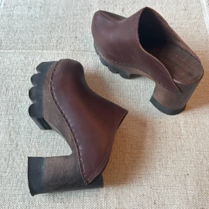 Bruna träklackade mules i skinn - Unika bruna mules med chunky träklack och plattformssula. Ovandel i brunt skinn med synliga sömmar och öppen häl. Sulan har coola räfflade detaljer för extra edge. Perfekta för dig som vill sticka ut med en retrovibe och statement-look.