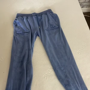 Mörkblå Juicy Couture M - Mysiga blå mjukisbyxor från Juicy Couture i velour med snörning i midjan och stora fickor fram och bak. Klassisk logga broderad på ena fickan. Perfekta för chill dagar eller när du vill ha en avslappnad look.
