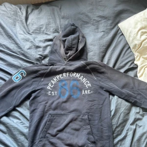 Mörkblå Peak Performance hoodie - Snygg mörkblå hoodie från Peak Performance med stor huva och tryck i vitt och blått på ryggen och ena ärmen. Klassisk känguruficka framtill och coola kontrastsömmar. Perfekt för chill eller sportig stil.