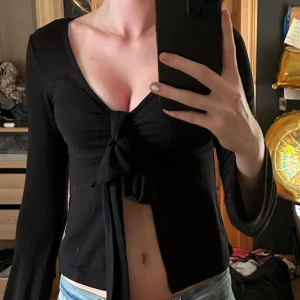 Svart omlottopp med knyt från Gina Tricot - Svart långärmad omlottopp från Gina Tricot i mjuk bomullsmix. Djup v-ringning och snygg knytning framtill som ger en croppad känsla. Perfekt att styla med jeans eller kjol för en trendig look.