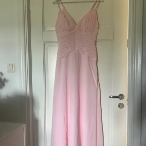 Ljusrosa långklänning med v-ringning - Superfin ljusrosa långklänning med dubbla smala axelband och v-ringning. Köptes second hand men användes aldrig. Klänningen har rynkade detaljer över byst och midja som ger en snygg siluett. Flödande kjol i tunt, lätt material – perfekt afton eller balklänning. 