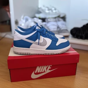 Nya Nike Dunk Low blå och vita sneakers - Nike Dunk Low sneakers i fräscha blå och vita färger med grå Swoosh. Klassisk låg modell med snörning och platt sula. Ovandel i skinn och syntet, perfekt för dig som gillar streetwear och vill sticka ut med en ikonisk siluett. Helt nya och kommer med original skolåda. 
