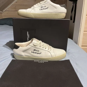 Saint Laurent sneakers vita - Säljer ett par vita sneakers från Saint Laurent med svart detalj på hälen och handskriven logga på sidan. Skorna har snörning och platt sula, perfekt för en clean och stilren look. Klassisk modell med rund tå och diskret branding.