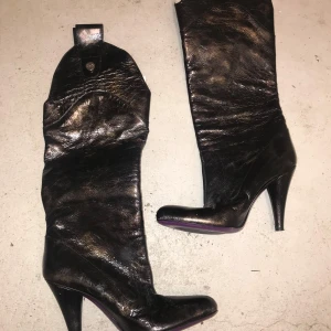 Svarta höga boots med klack - Säljer ett par svarta boots i glansigt skinn med hög klack och rund tå. Stövlarna har dekorativ knappdetalj upptill och lila sula undertill. Perfekta för dig som vill sticka ut med din stil.