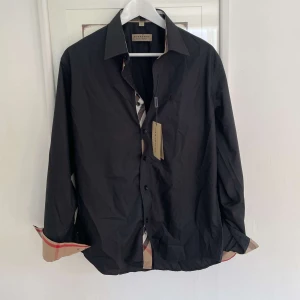 Svart skjorta från Burberry - Stilren svart skjorta från Burberry med klassiska rutiga detaljer på insidan av kragen och ärmsluten. Skjortan har knappar framtill och lång ärm. Perfekt för dig som gillar en tidlös look med en twist.