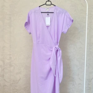 Ljuslila omlottklänning ny  - Säljer en ljuslila omlottklänning med knytband i midjan och korta ärmar. Klänningen har en v-ringad hals och är i en mjuk, lätt kvalitet. Perfekt för vår och sommar!