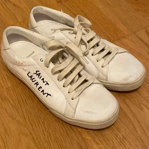 Vita sneakers från Saint Laurent - Säljer ett par klassiska vita sneakers från Saint Laurent med svart handskriven logga på sidan. Skorna har rund tå, platt sula och snörning. Perfekta för en stilren och avslappnad look.