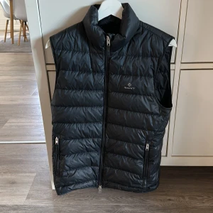 Gant dunväst  - Säljer en svart dunväst från Gant med hög krage och två fickor med dragkedja. Västen har quiltad design och stängs med dragkedja framtill. Perfekt för lager-på-lager under kyliga dagar. Västen är i bra skick och endast använd i ett år. nypris 2000kr Storlek XS