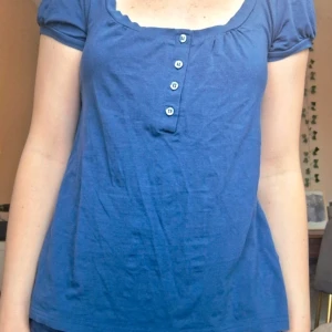 Blå kortärmad topp med knappar - Säljer en blå topp med korta ärmar och rundad urringning. Toppen har tre knappar framtill och är gjord i ett mjukt bomullsmaterial. Perfekt till vardags och enkel att matcha med jeans eller kjol. Tröjan köptes på vinted och bilden är  lånad från säljaren.